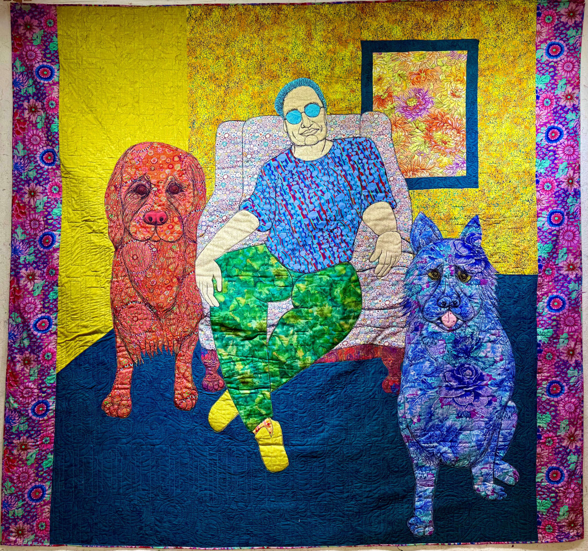 A colorful applique quilt of various floral fabrics. The center of the quilt shows a man in glasses seated in an armchair with an orange dog on his right and a blue dog on his right.